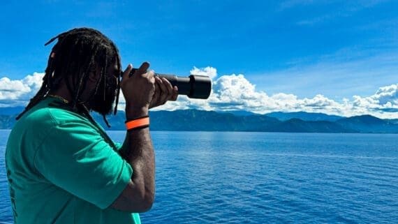 Travel creator photographing the ocean from a ferry in the Philippines using a telephoto camera lens, with blue skies, mountains, and open water in the background.