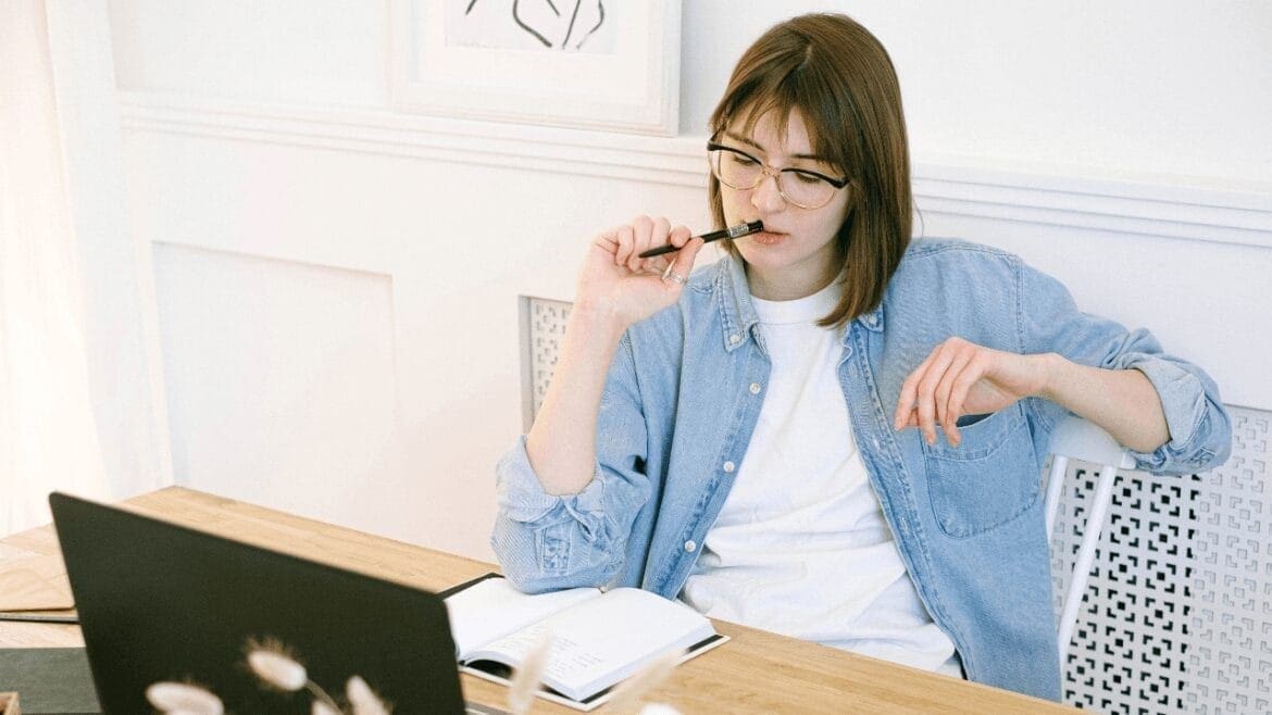 Young woman in glasses and denim shirt sitting at a desk with a laptop and notebook, holding a pen near her mouth thoughtfully.