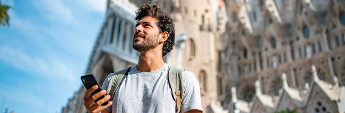 A man with a backpack looking up while holding a smartphone, with a historic cathedral in the background.
