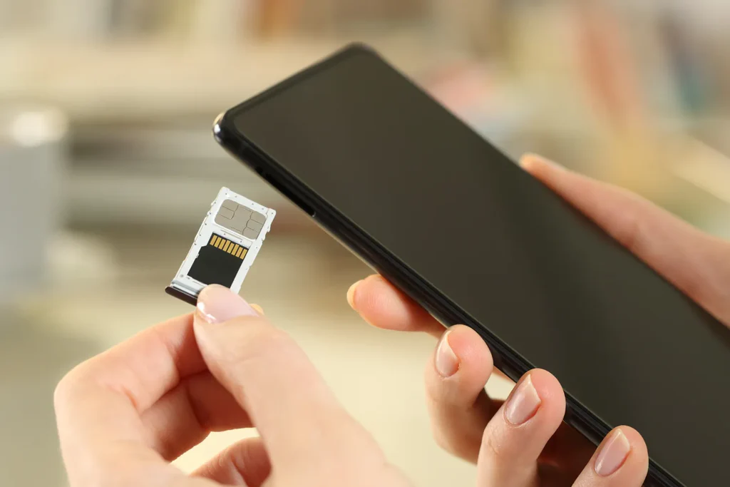 A person inserts a SIM card into a smartphone, holding the SIM tray with one hand and the phone with the other, against a blurred indoor background.