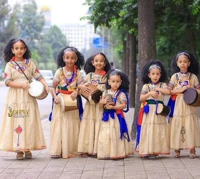 Seven young girls in beige dresses with blue and red trim stand in a row on a sidewalk, each holding a drum. They have matching braided hairstyles and headbands, with greenery and trees behind them. The scene exudes community, tradition, and youthful joy.