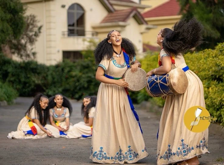 Young women in cream dresses with blue embroidery perform Ashenda: two center-stage girls cheer while beating drums, others sit and smile in the background, all sharing a moment of dance and sisterhood in a sunlit outdoor setting during Ashenda season.