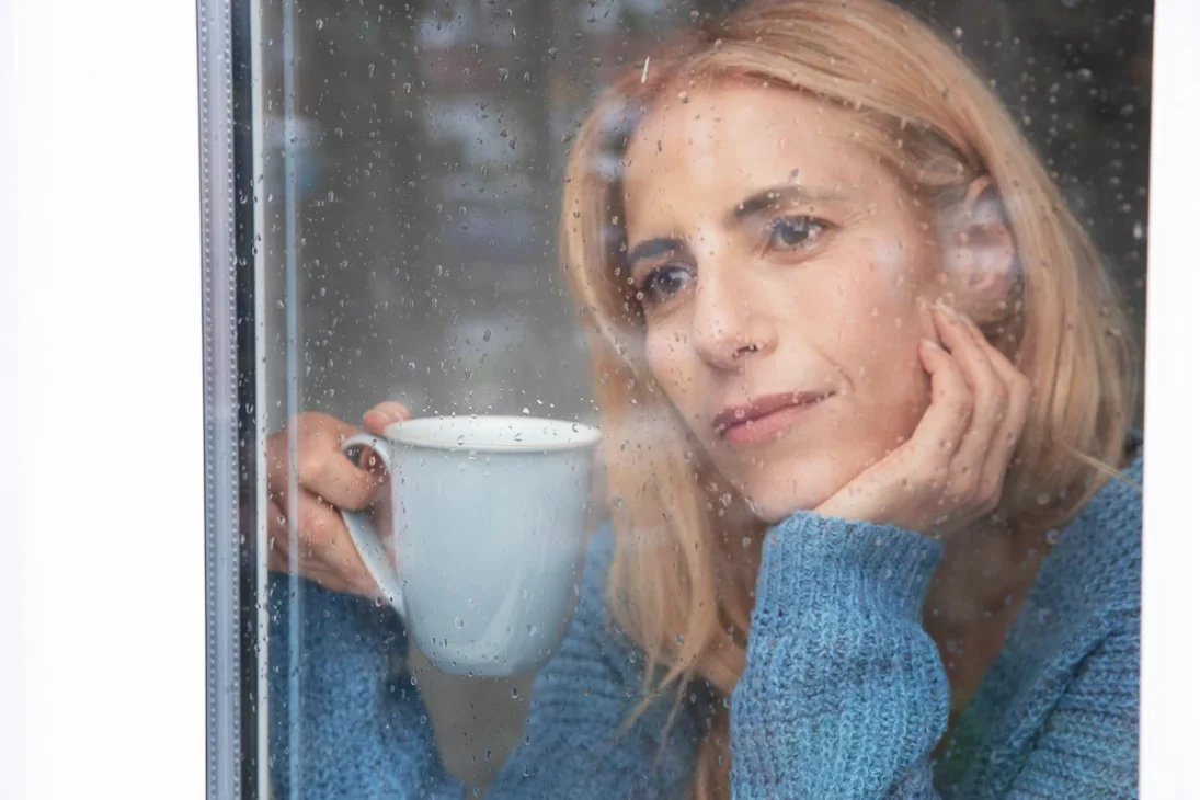 Woman holding a cup, looking out the window with a neutral expression, reflecting on the weather.