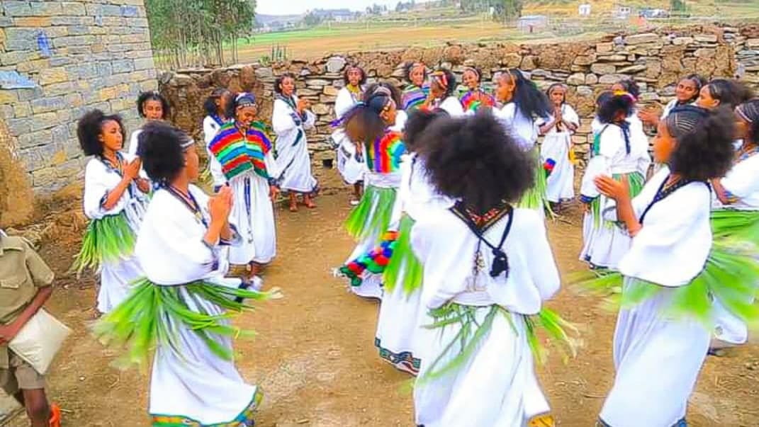 Group of young women wearing white dresses with green leaves on their waist form a circular dance outdoors, clapping and moving to the rhythm. A stone wall and rural background frame the scene, conveying a festive community event.