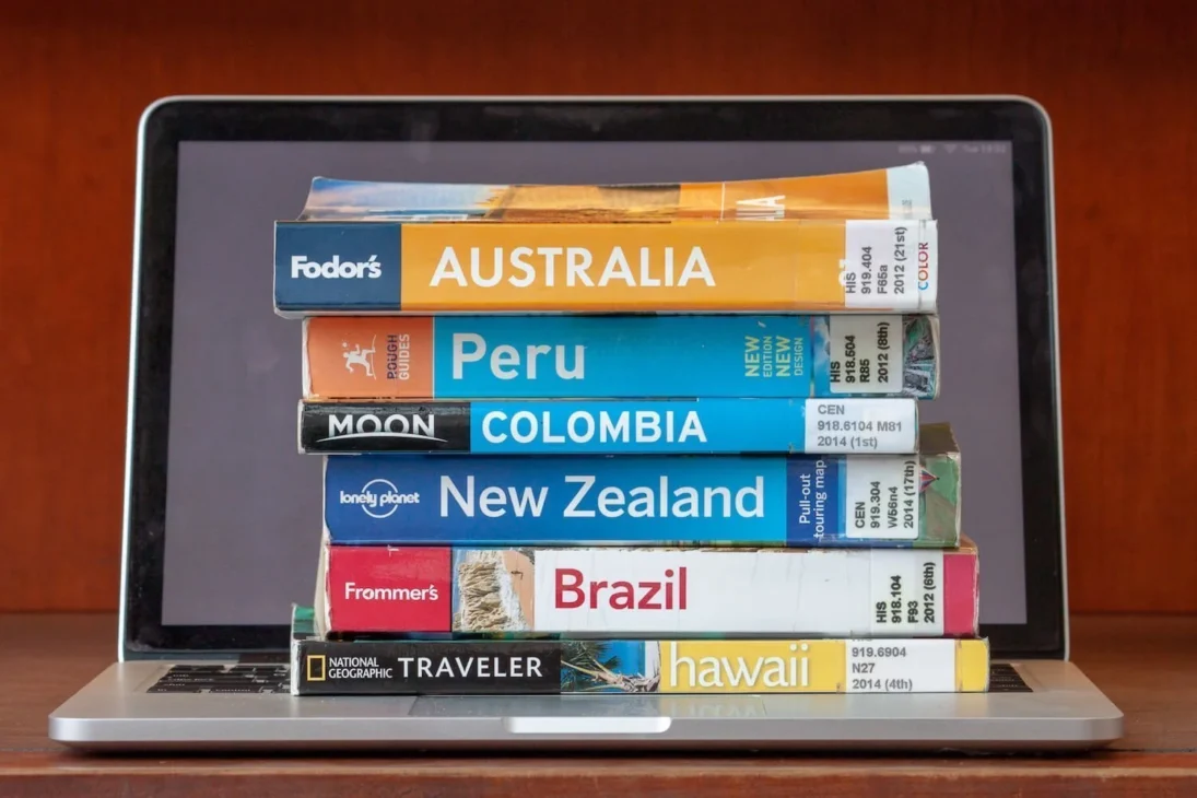 A stack of colorful print travel guidebooks for various countries (Australia, Peru, Colombia, New Zealand, Brazil, and Hawaii) resting directly on the keyboard of a silver laptop.