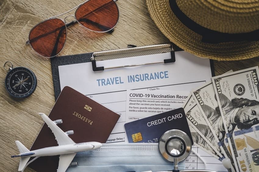 Flat lay image of key travel essentials on a wooden table: a passport, cash, a credit card, a toy airplane, a stethoscope, and a document clearly labeled "TRAVEL INSURANCE." This illustrates the critical importance of verifying policy legitimacy and organizing documents before departure.