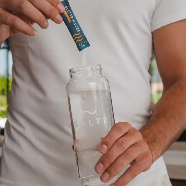 A person is pouring powdered electrolytes into a clear water bottle to support hydration and help recover after long flight.